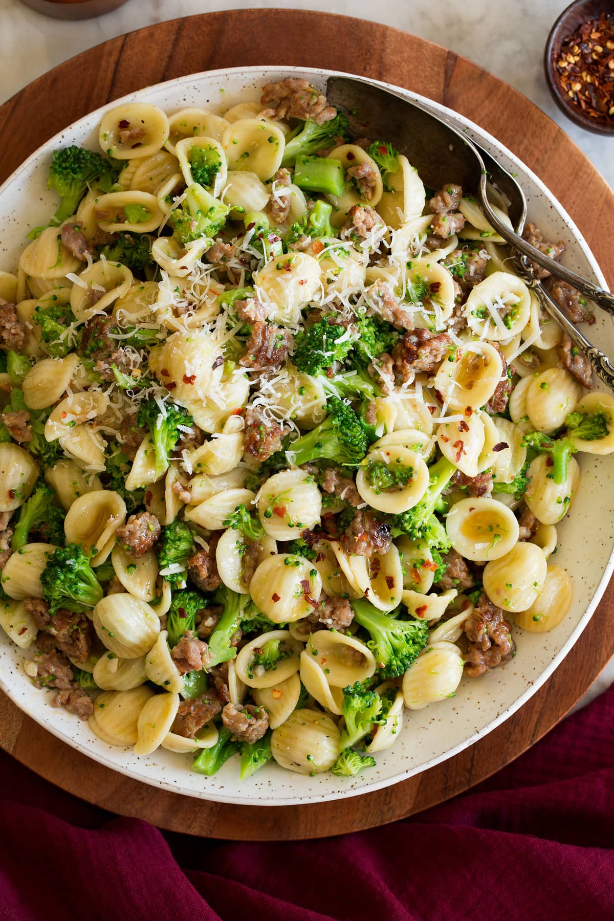Photo: Orecchiette pasta with sausage and broccoli in a white serving bowl set over a wooden platter on a marble surface. It is shown from above.