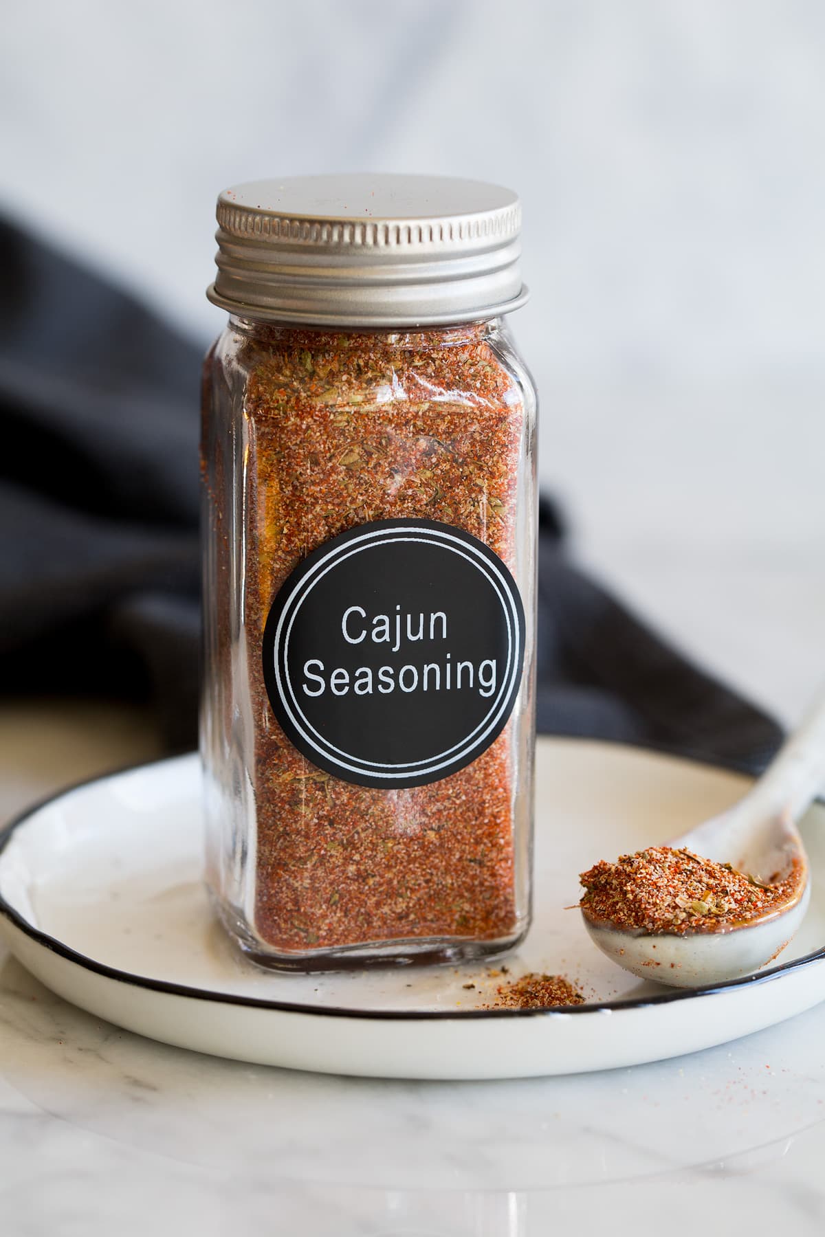 Photo: Homemade cajun seasoning blend in a glass spice jar on a small plate with a spoon to the side and a dark napkin in the background.