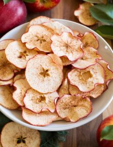 Apple chips shown in a white bowl on a wooden tabletop with fresh apples and leaves surrounding them.