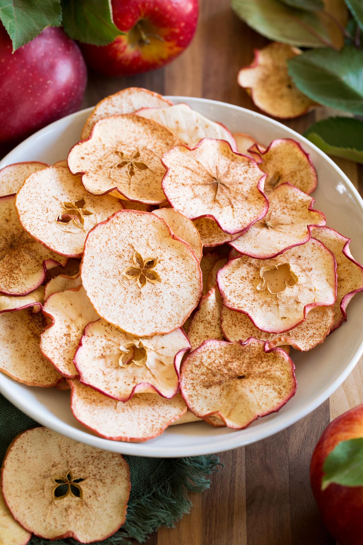 Apple chips shown in a white bowl on a wooden tabletop with fresh apples and leaves surrounding them.