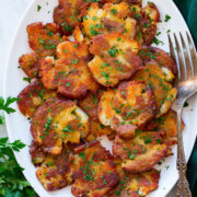Smashed red potatoes shown piled on a white oval platter. They are shown from above on a white marble surface with a green cloth and parsley to the sides.