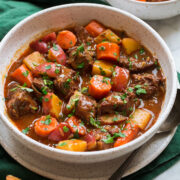 Beef Stew shown in a white serving bowl over a green cloth. Bread is shown to the side as a serving suggestion.