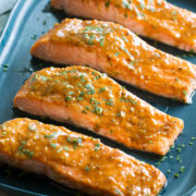 Photo of four salmon fillets coated with a brown sugar glaze resting on a blue rectangular platter on a white marble surface.