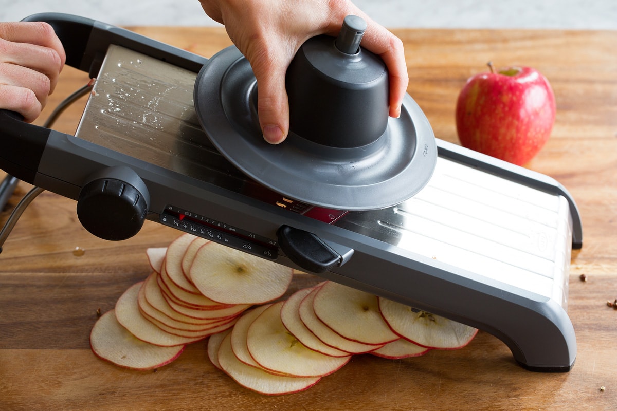 Apples being sliced with a mandoline into thin slices over a wooden cutting board.