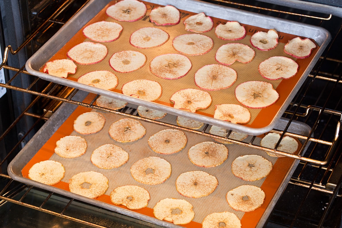 Apple slices being baked in an oven on two baking sheets set on two oven racks.