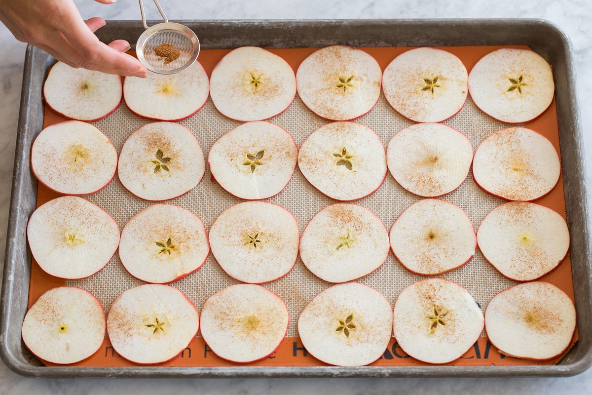 Thin apple slices on a silicone baking mat lined baking sheet being dusted with cinnamon.