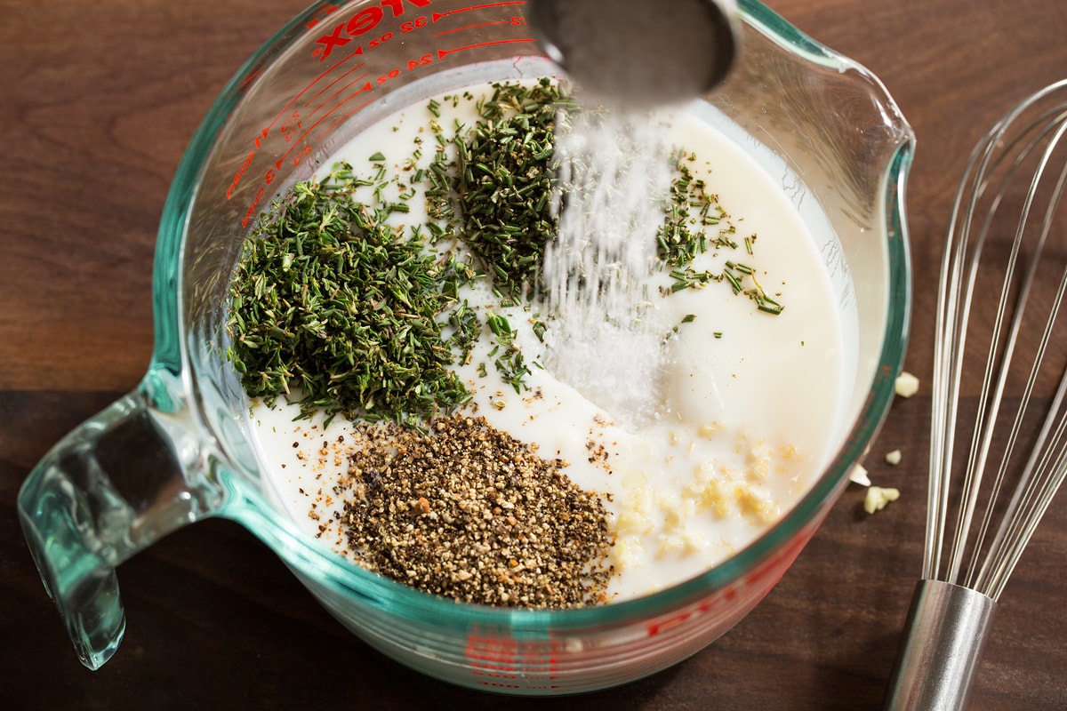 Buttermilk, salt, herbs, garlic and pepper shown before mixing in a glass measuring cup.