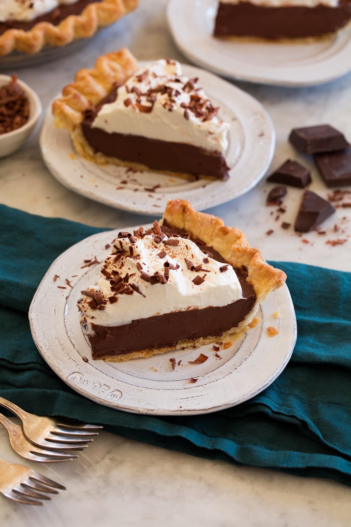 Two slices of chocolate cream pie on small white dessert plates set over a blue cloth on a marble surface with chocolate chunks and curls in the backround.