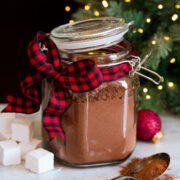 Homemade hot chocolate mix in a glass canister with a buffalo check red and black ribbon. A Christmas tree is in the background.