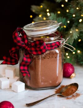 Homemade hot chocolate mix in a glass canister with a buffalo check red and black ribbon. A Christmas tree is in the background.