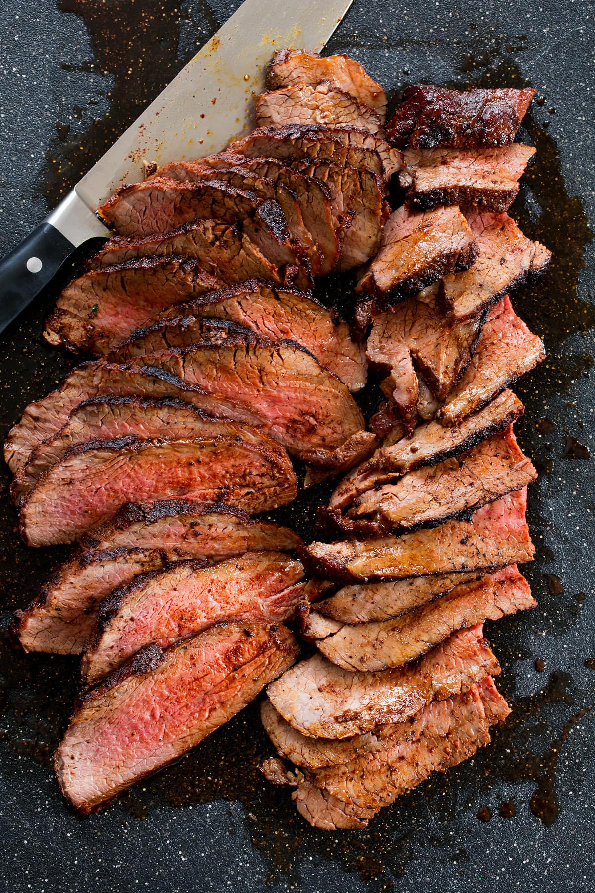 Overhead photo of tri tip being cut on a black cutting board with a chefs knife to the side.