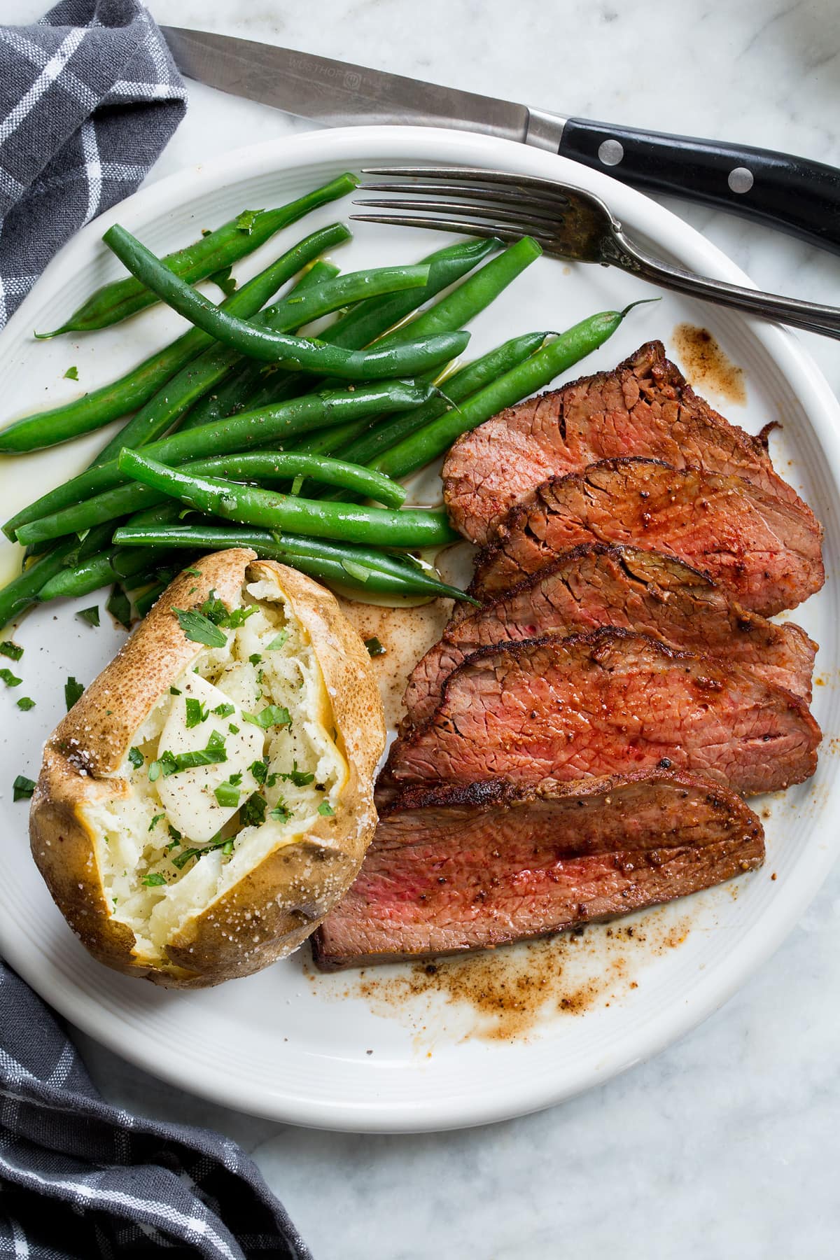 Roasted tri tip on a white plate shown with serving suggestion of baked potato and steamed green beans.