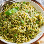Homemade pesto tossed with spaghetti pasta in a large white serving bowl set over a wooden platter.