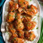 Slow cooker chicken thighs with sauce shown served over white rice on a white oval platter. Platter is on a marble surface with a blue cloth and green onions to the side.