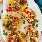 Breakfast tacos made with flour tortillas, chorizo, bacon, eggs, cheese, avocado and pico de gallo shown on a white oval platter from above.