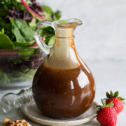 Balsamic vinaigrette in a glass salad dressing jar set over a small plate. A green salad is shown in the background.