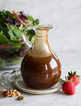 Balsamic vinaigrette in a glass salad dressing jar set over a small plate. A green salad is shown in the background.