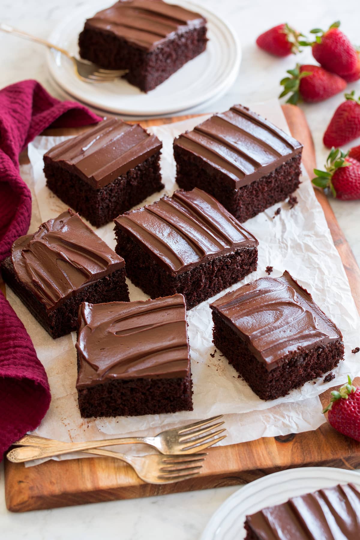 Square slices of homemade chocolate cake shown on a wooden serving board with parchment paper. Strawberries are shown as a serving suggestion to the side.