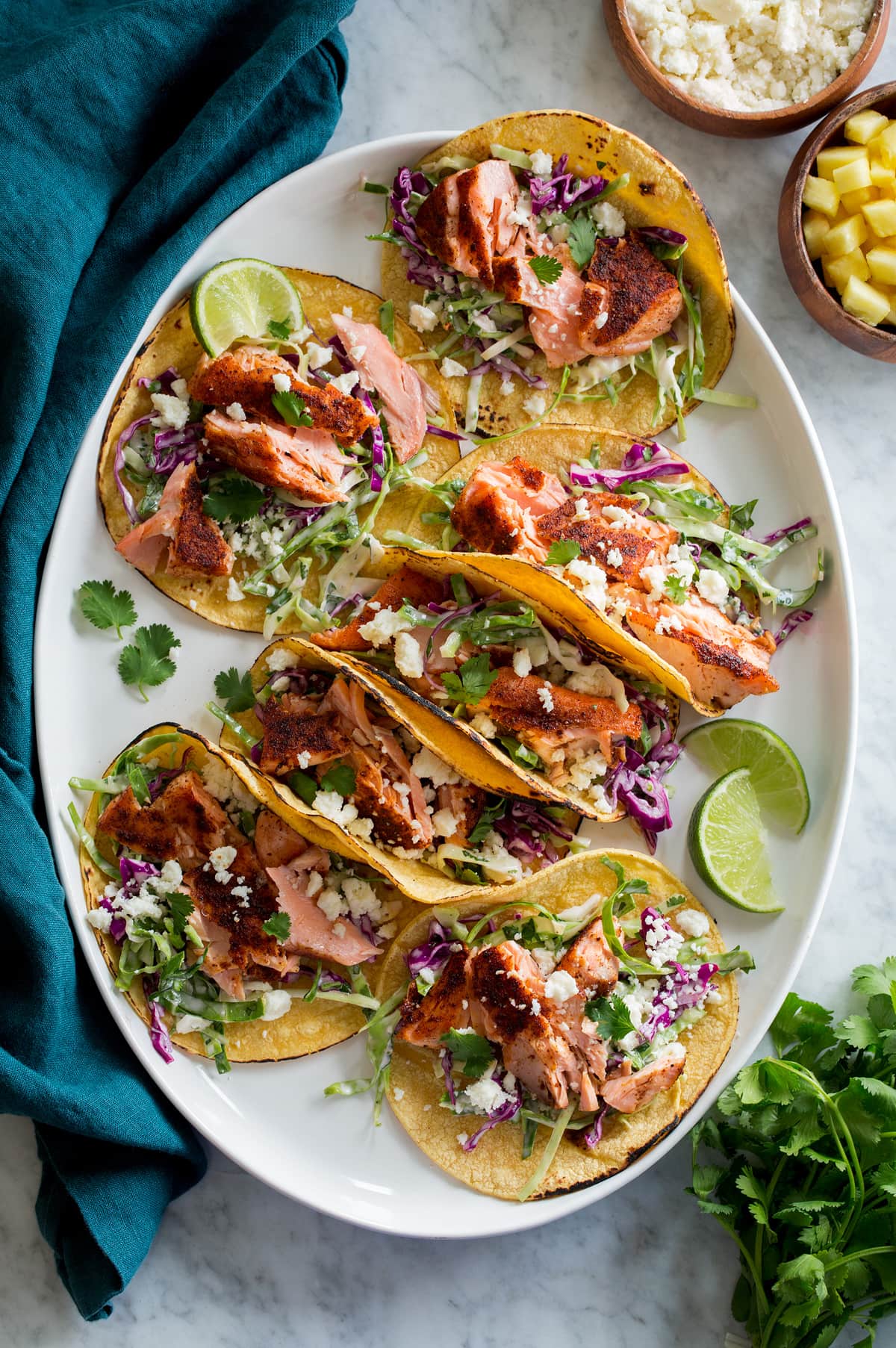 Salmon Tacos shown on a white oval platter on a marble surface. Salmon tacos a layered with corn tortillas, fish taco slaw, seasoned pan seared salmon, and queso fresco cheese. 