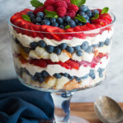 Layered trifle in a glass trifle dish. It includes cubes of angel food cake, blueberries, strawberries, raspberries and a cream cheese whipped cream topping.