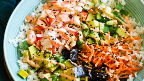 California roll sushi bowl shown in a blue bowl from a side angle. Bowl is resting on a wooden plate over a marble surface.