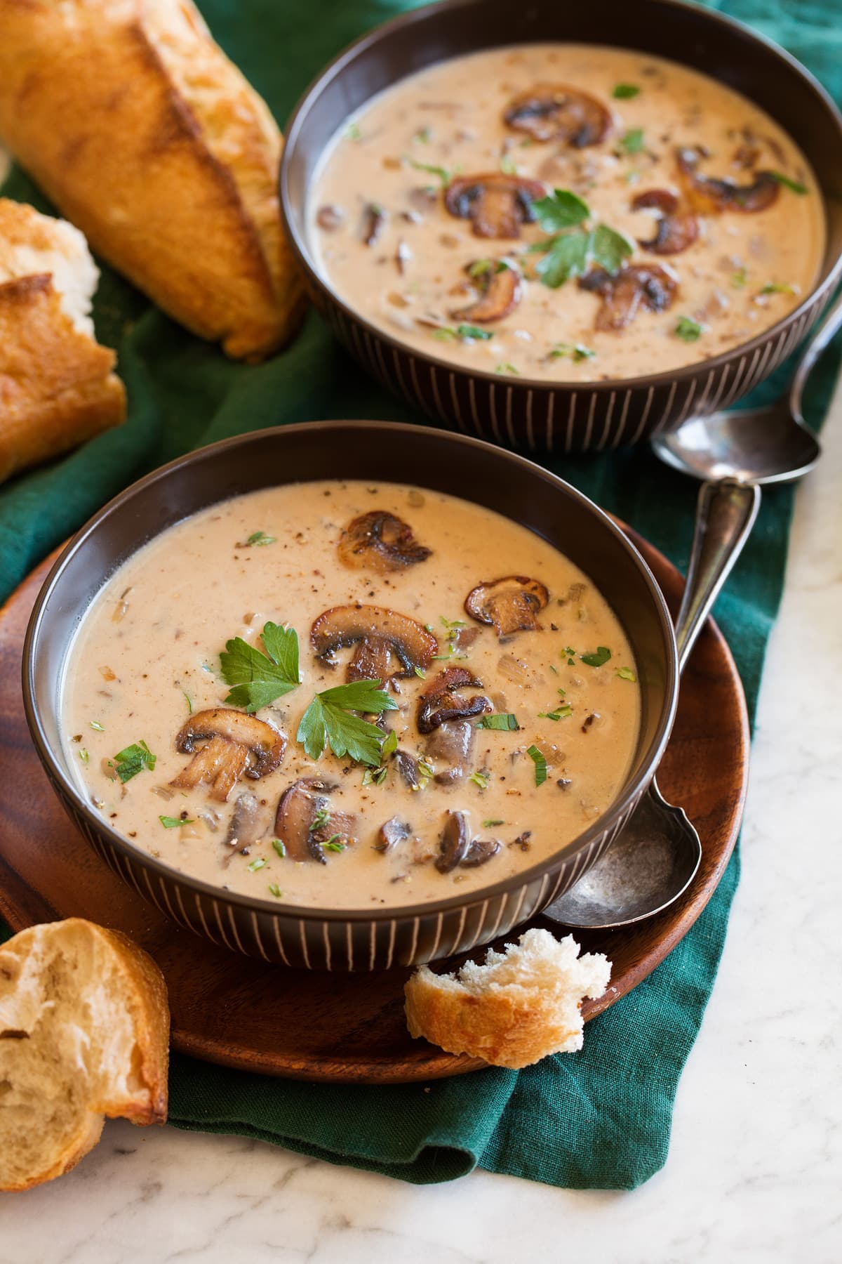 Two servings of mushroom soup in brown bowls sitting on a green cloth on a marble surface.