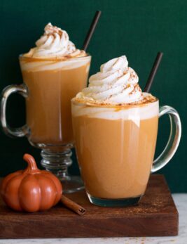 Pumpkin spice latte. Shows two servings in glass mugs one a wooden plate set over a white marble surface with a green background.