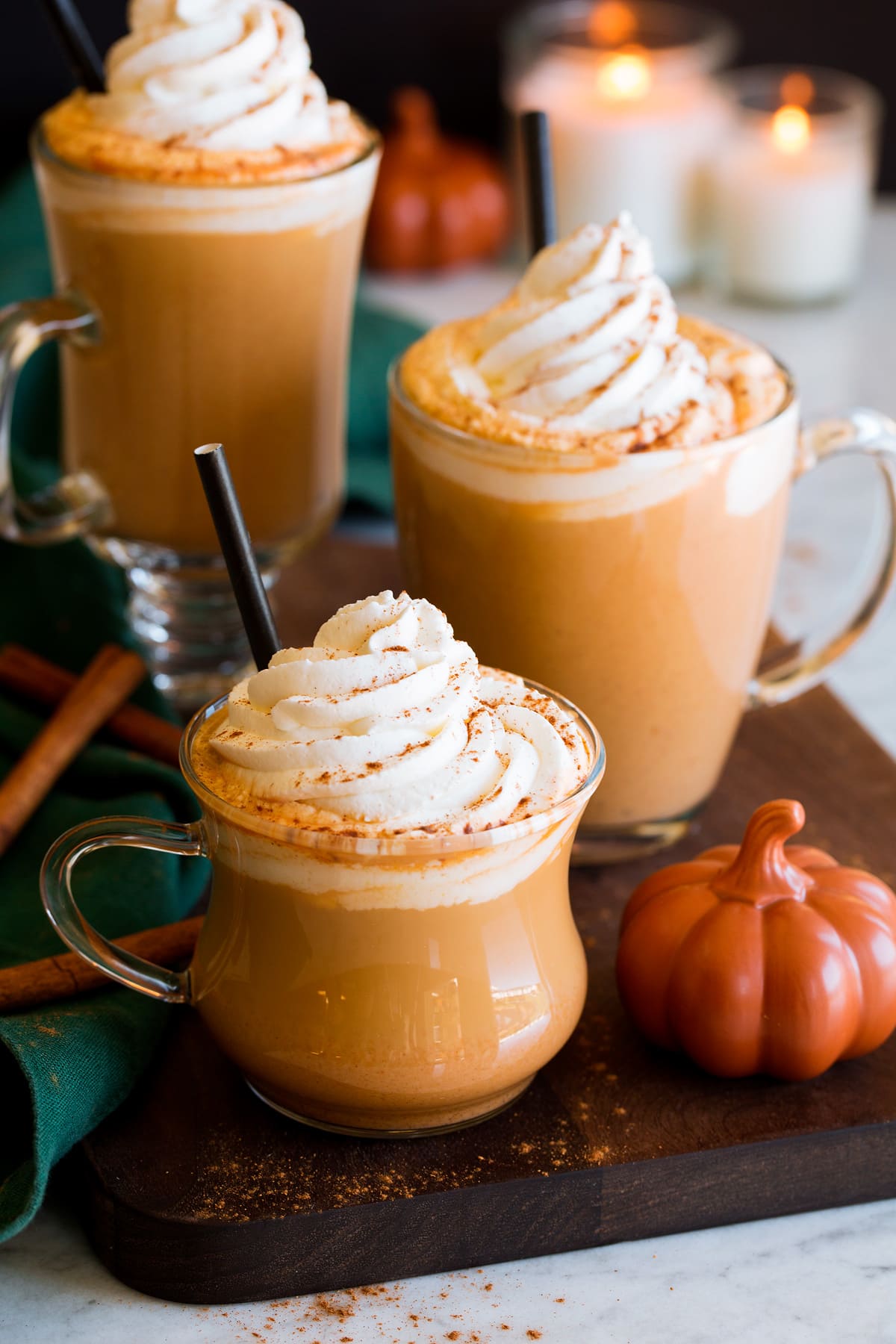 Three pumkpin spice lattes with a decorative pumpkin to the side, cinnamon sticks and candles in the background. Lattes are topped with whipped cream and pumpkin spice.