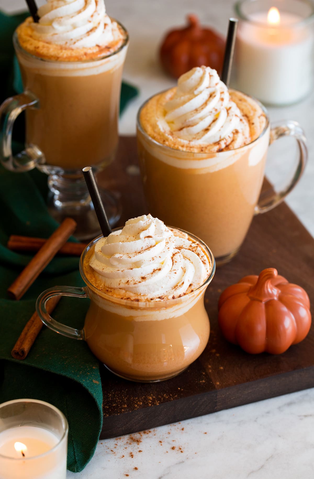 Three pumkpin spice lattes with a decorative pumpkin to the side, cinnamon sticks and candles in the background. Lattes are topped with whipped cream and pumpkin spice.