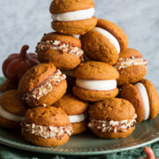 Pumpkin Whoopie Pies stacked on a large plate. They are filled with cream cheese frosting and some are covered with chopped pecans.