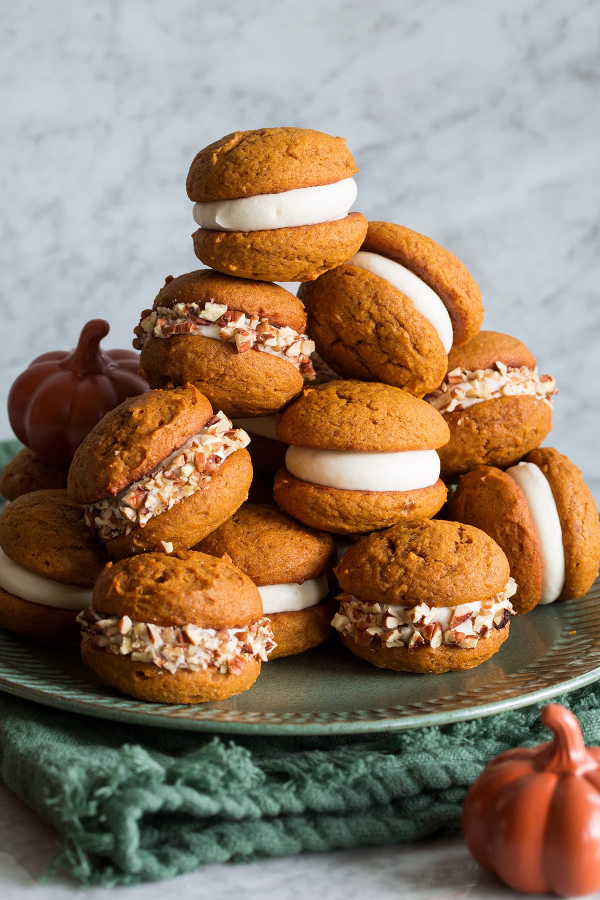 Pumpkin Whoopie Pies stacked on a large plate. They are filled with cream cheese frosting and some are covered with chopped pecans.