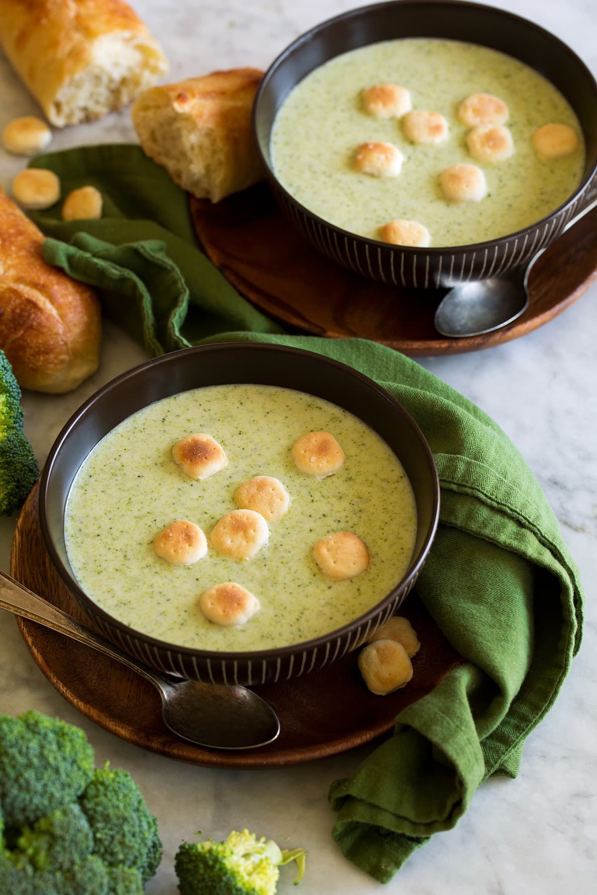 Creme de sopa de brócolis em tigelas marrons com bolachas em cima.  Um pano verde é mostrado ao lado e pão baguete como sugestão de servir.