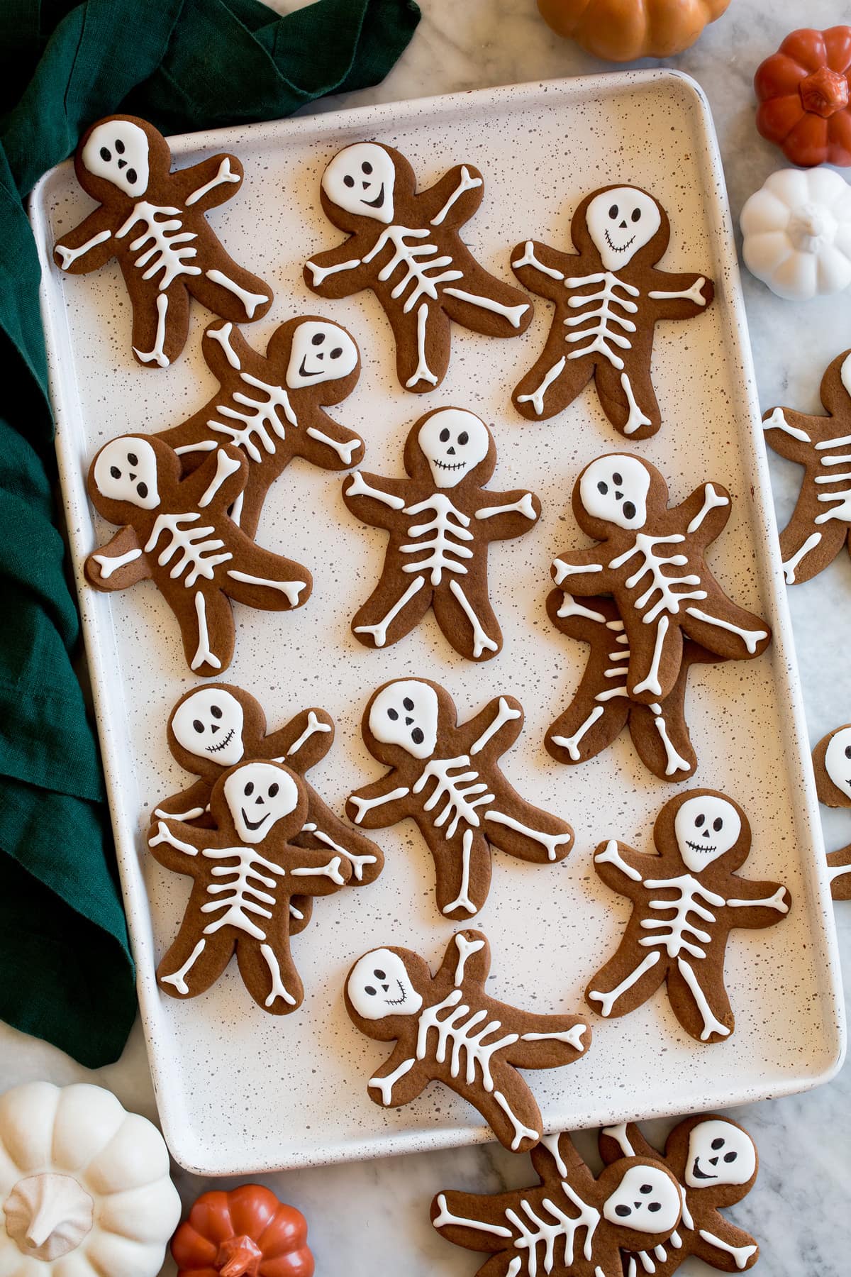 Skeleton gingerbread cookies on a white baking sheet with a green cloth and decorative pumpkins to the side.