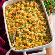 Cornbread stuffing shown in a white rectangular baking dish over a wooden platter with herbs, cranberries and a red cloth to the side.