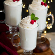 Three mugs of white hot chocolate on a wooden tray with Christmas lights in the background.