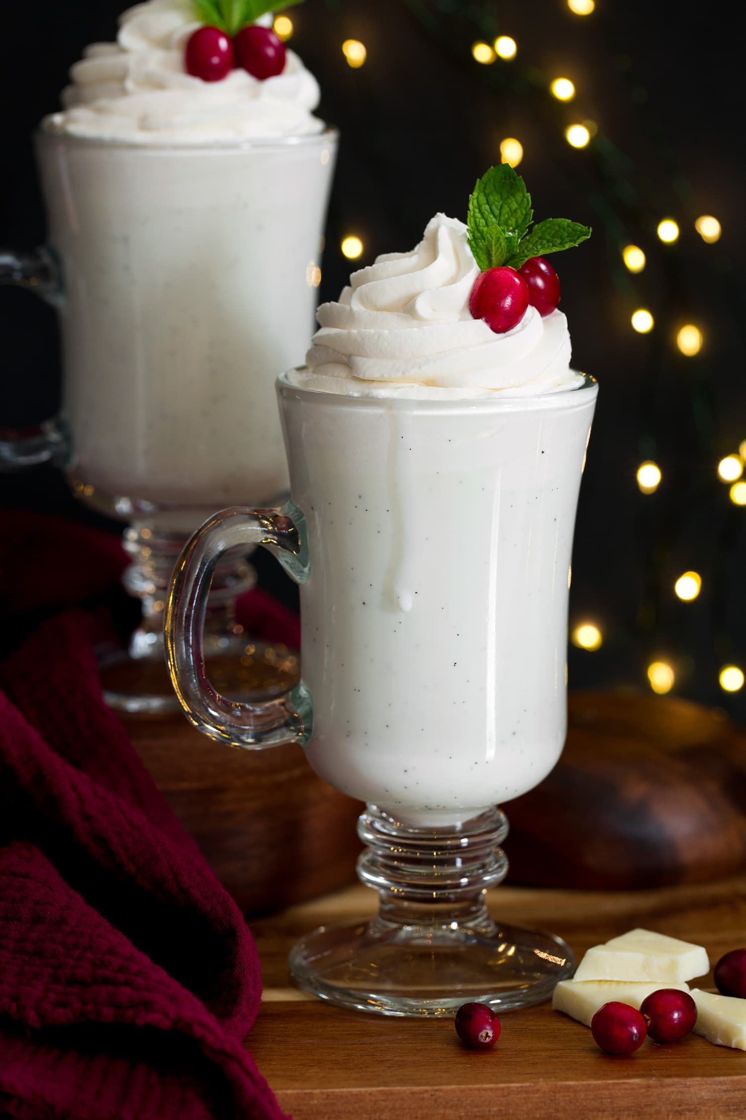Close up photo of tall glass of white hot chocolate topped with whipped cream, cranberries and mint.