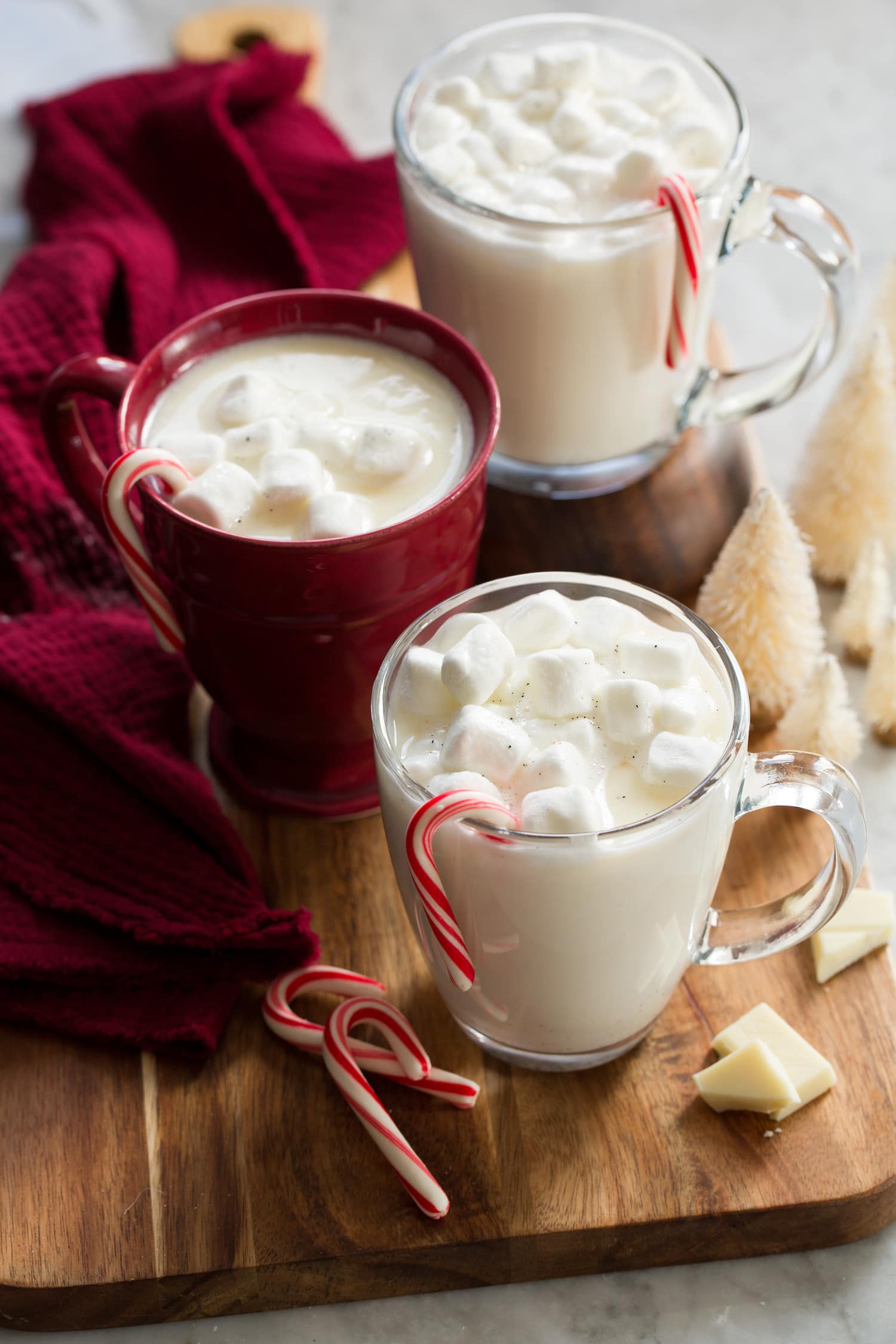 Three cups of white hot chocolate topped with marshmallows and a peppermint candy cane hanging on each mug.