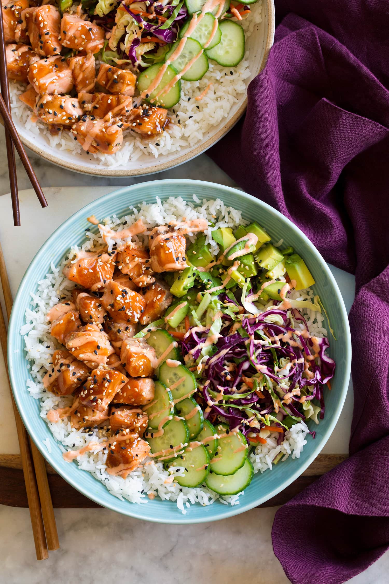 Layered into a blue bowl is white rice, glazed salmon, cucumber, asian cabbage slaw, avocado, and sesame seeds.