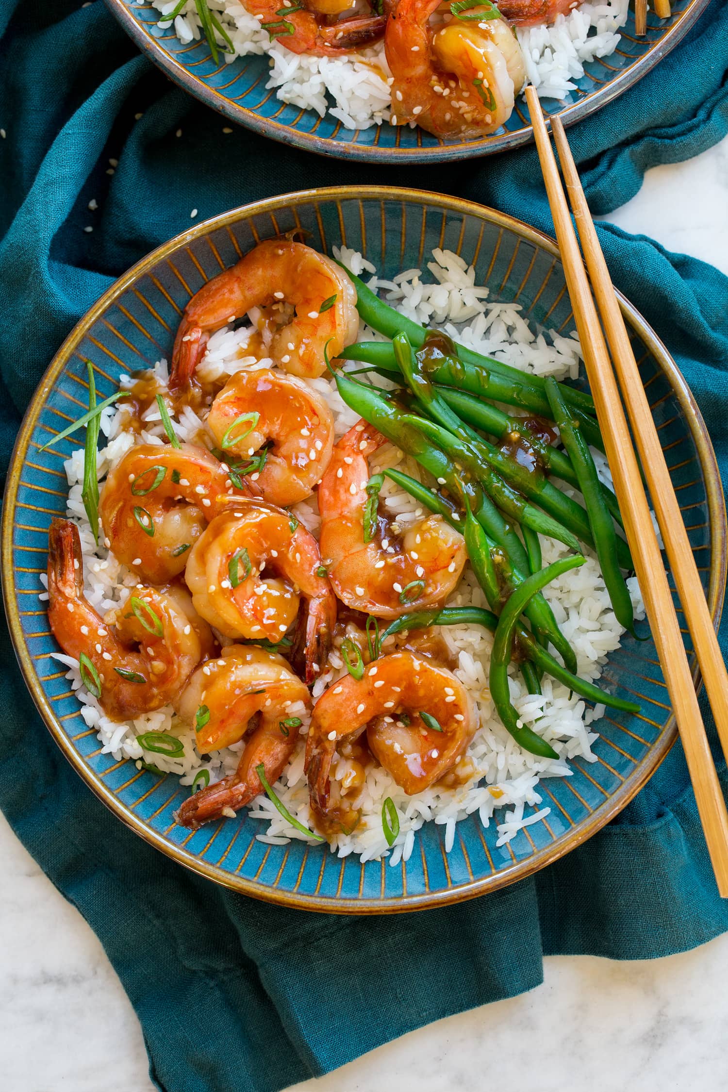 Teriyaki shrimp in a blue bowl with rice, green beens and green onions. Shown with a blue cloth and chopsticks.
