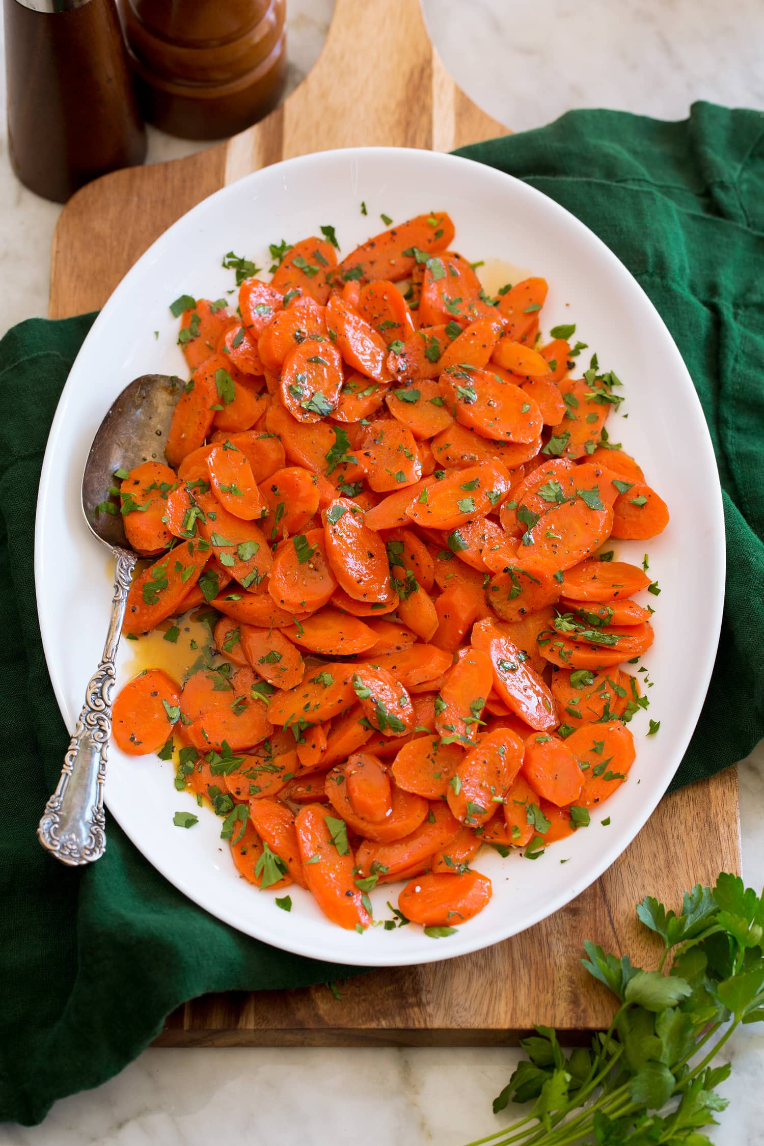 Glazed carrots shown served on a white oval platter set over a wooden tray and a green cloth.