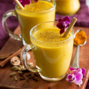 Golden milk shown in glass mugs on a wooden tray with spices and flowers to the side.