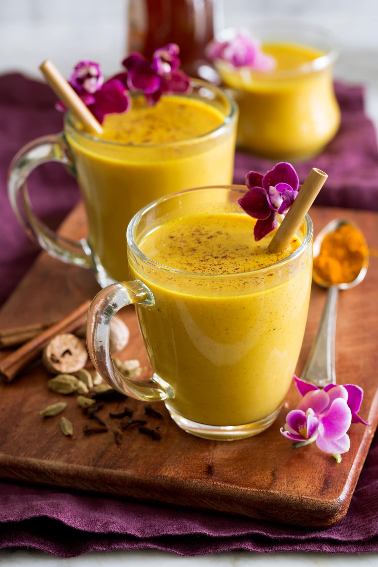 Golden milk shown in glass mugs on a wooden tray with spices and flowers to the side.
