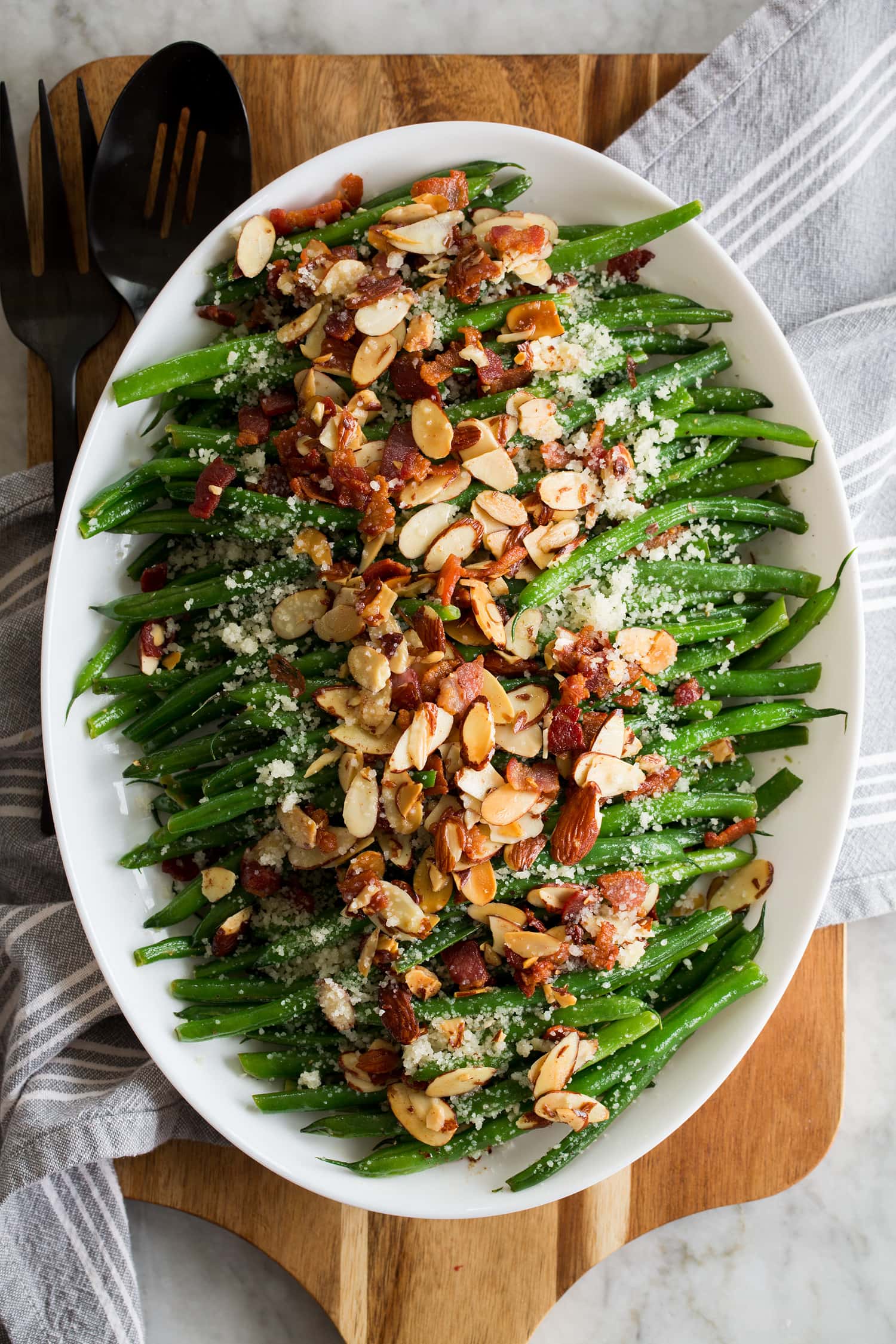Beautiful Green Bean Almondine. Sauteed green beans are shown lined up in a row on a platter with toasted almonds, crumbled fried bacon and grated parmesan are sprinkled over the top.