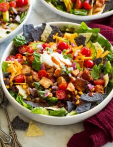 Chicken taco salad shown with blue and yellow tortilla chips, corn, avocado, tomatoes, lettuce, chicken, cilantro, cheese and sour cream.