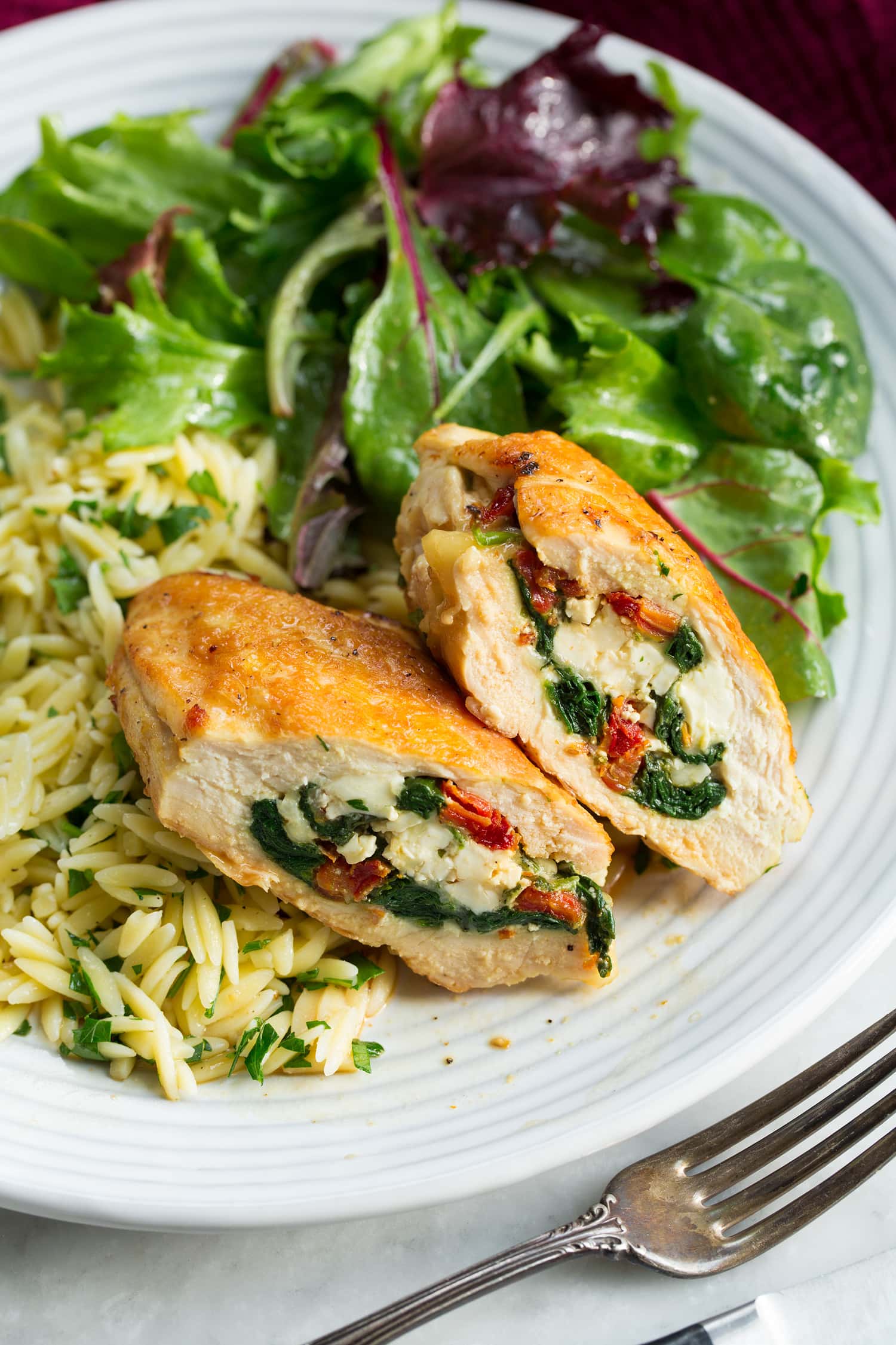Two sliced stuffed chicken breasts halves shown with serving suggestions of side salad and lemon orzo.