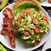 Avocado chicken salad shown on wheat bread with lettuce leaves and sliced grape tomatoes.