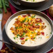 Serving of baked potato soup in a gray bowl over a gray plate. It is topped with cheddar cheese, fried potato skins, bacon and green onions.
