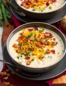 Serving of baked potato soup in a gray bowl over a gray plate. It is topped with cheddar cheese, fried potato skins, bacon and green onions.