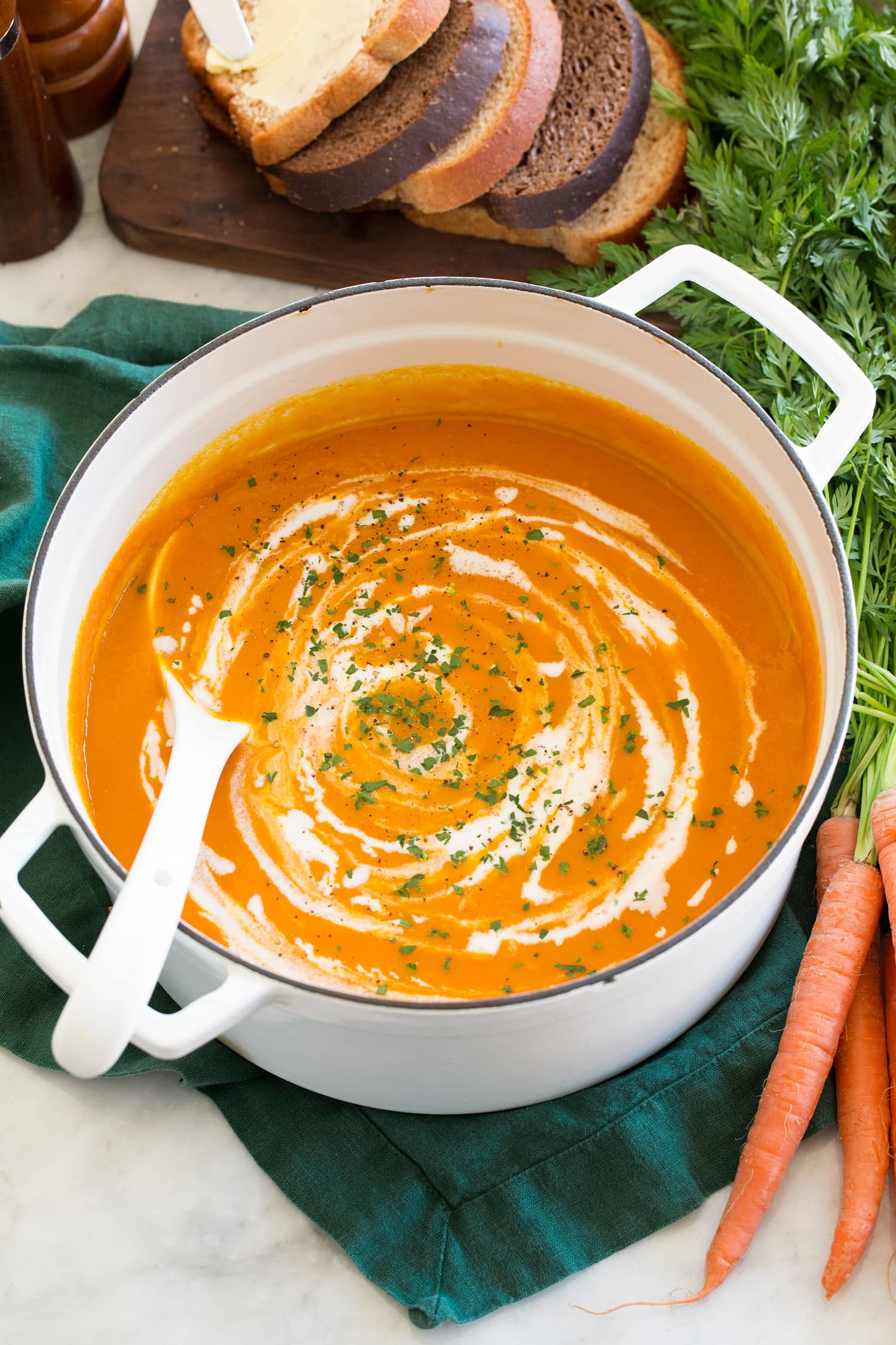 Carrot soup shown in a white enameled pot over a green cloth with fresh carrots to the side.