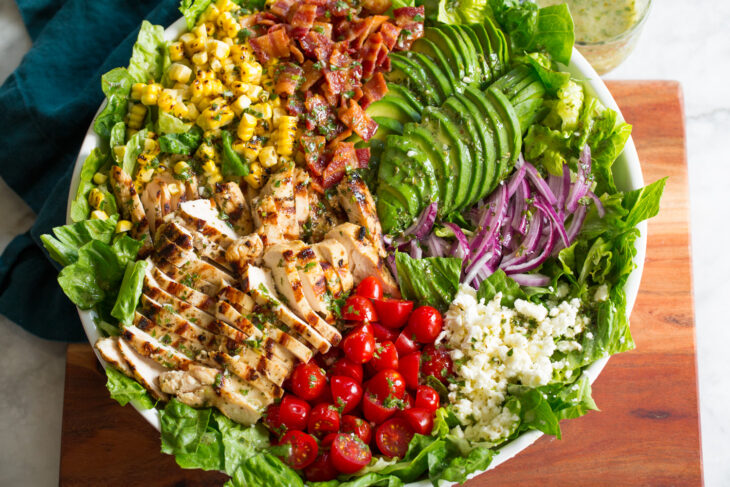 A large salad bowl filled with chopped ingredients, including grilled chicken, cherry tomatoes, avocado slices, diced bacon, sweet corn, red onion, crumbled feta cheese, and a green leafy base. A small cup of dressing is visible in the background.